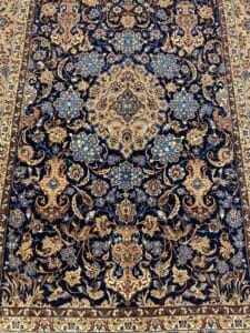 Azerbaijan Tappeti Milano Tappeto persiano ricco di dettagli con intricati motivi floreali nei toni del blu, del beige e del marrone, caratterizzato da un medaglione centrale simmetrico e da bordi ornati. Nell'angolo in alto a destra compare il logo di una foglia colorata.