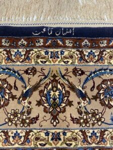 Azerbaijan Tappeti Milano Primo piano di un tappeto persiano con intricati disegni floreali in blu, marrone e beige. Il bordo superiore presenta frange color crema, un bordo blu con scritte persiane e un logo floreale colorato nell'angolo in alto a destra.