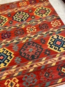 Azerbaijan Tappeti Milano Un tappeto colorato con motivi geometrici nei toni del rosso, giallo, blu e verde, posato su un pavimento piastrellato. Il tappeto presenta motivi a rombi e croci, con un logo a forma di foglia multicolore nell'angolo.