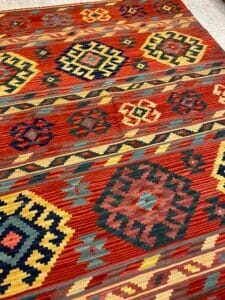 Azerbaijan Tappeti Milano Un tappeto colorato a tessitura piatta con motivi geometrici in rosso, giallo, blu, nero e verde, con disegni a rombi e a zig-zag. Nell'angolo in alto a destra compare il logo di una farfalla multicolore.