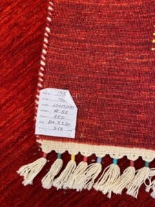 Azerbaijan Tappeti Milano Primo piano di un tappeto rosso con un bordo bianco sfrangiato. Un'etichetta mostra dettagli come dimensione e colore. Un logo floreale colorato è visibile nell'angolo.