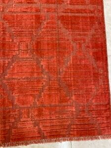 Azerbaijan Tappeti Milano Tappeto rosso testurizzato con motivi geometrici e un logo colorato in un angolo, posizionato su un pavimento piastrellato chiaro.