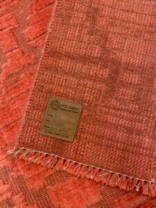 Azerbaijan Tappeti Milano Un tappeto testurizzato rosso e arancione con un'etichetta in legno che ne mostra il codice, la misura e altri dettagli. Un logo colorato si trova nell'angolo in alto a destra.