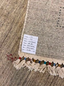 Azerbaijan Tappeti Milano Primo piano di un tappeto intrecciato che mostra un'etichetta con dettagli come numero dell'articolo, colore, dimensione e prezzo. Il tappeto ha nappe multicolori e un sottile motivo geometrico.