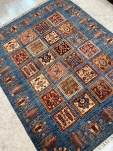 Azerbaijan Tappeti Milano Un tappeto multicolore con un motivo a griglia di motivi quadrati su sfondo blu, caratterizzato da disegni rossi, marroni e beige, adagiato su un pavimento piastrellato.