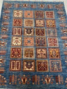 Azerbaijan Tappeti Milano Un tappeto vivace presenta una griglia di intricati motivi quadrati nei toni del rosso, del blu e del beige su uno sfondo blu, con motivi floreali decorativi lungo i bordi.