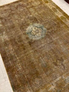 Azerbaijan Tappeti Milano Un tappeto sbiadito con un motivo a medaglione centrale, adagiato su un pavimento piastrellato. Una foglia multicolore e un logo stilizzato "A" sono visibili nell'angolo.