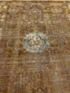 Azerbaijan Tappeti Milano Un tappeto rettangolare, usurato, con un motivo floreale sbiadito in toni verdi e marroni tenui. Un logo colorato è nell'angolo in alto a destra.