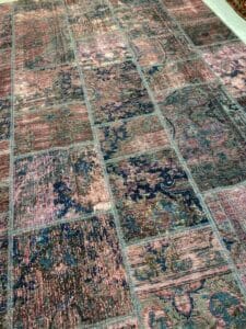 Azerbaijan Tappeti Milano Un grande tappeto patchwork con motivi intricati nelle tonalità del rosa, blu e grigio, caratterizzato da un logo colorato nell'angolo con la lettera "A" e un motivo a foglie multicolori.