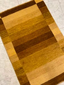 Azerbaijan Tappeti Milano Un tappeto rettangolare con un motivo a scacchi in tonalità di marrone e beige, esposto su un pavimento di piastrelle chiare. C'è un logo colorato nell'angolo in alto a destra.