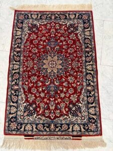 Azerbaijan Tappeti Milano Tappeto persiano rettangolare con centro rosso, intricati motivi floreali in crema, blu e beige, bordo blu scuro e frange su entrambe le estremità. Lungo il bordo inferiore è presente una scritta persiana. Il tappeto si trova su un pavimento di piastrelle chiare.