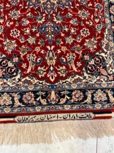 Azerbaijan Tappeti Milano Un tappeto persiano dettagliato con un motivo floreale rosso e blu, frange color crema e un bordo color crema con scritte persiane. Una piccola girandola colorata si trova nell'angolo superiore destro del tappeto.