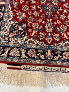 Azerbaijan Tappeti Milano Primo piano di un tappeto persiano con motivo floreale rosso e blu, frange beige e scritte persiane tessute lungo il bordo. Il tappeto poggia su una superficie chiara. Nell'angolo in alto a destra è presente un logo a forma di fiore colorato.