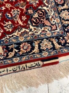 Azerbaijan Tappeti Milano Primo piano di un tappeto persiano tradizionale con intricati motivi floreali nei colori rosso, blu e crema, con frange lungo il bordo e scritte decorative in farsi tessute vicino al bordo.