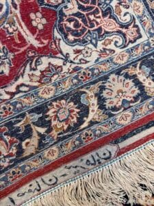 Azerbaijan Tappeti Milano Primo piano di un tappeto decorato con motivi floreali rossi, blu e beige, frange color crema e una sezione di scrittura persiana lungo il bordo. Un logo colorato appare nell'angolo in alto a destra.