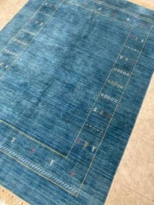 Azerbaijan Tappeti Milano Un tappeto blu con un semplice motivo geometrico a bordatura con piccoli motivi decorativi. Il tappeto è posizionato su un pavimento piastrellato di colore chiaro.