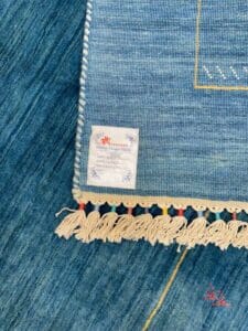 Azerbaijan Tappeti Milano Una sezione di un tappeto blu con una frangia decorativa e un'etichetta. L'angolo presenta un logo multicolore con un fiore stilizzato e un testo sottostante.