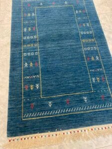 Azerbaijan Tappeti Milano Un tappeto rettangolare blu con motivi geometrici e floreali in bianco, rosso e giallo. Un logo ricamato multicolore è nell'angolo in alto a destra. Nappe con frange sono sul bordo inferiore.