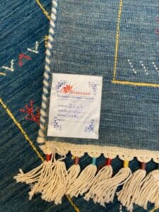 Azerbaijan Tappeti Milano Primo piano di un tappeto blu intrecciato con un'etichetta decorativa con motivi floreali, che riporta informazioni dettagliate su restauro e pulizia. Nappe multicolori ne delineano il bordo.