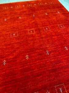 Azerbaijan Tappeti Milano Un tappeto rosso con piccoli motivi geometrici e vegetali. Un logo a forma di foglia multicolore è nell'angolo in alto a destra.