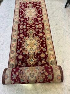 Azerbaijan Tappeti Milano Un tappeto runner rosso e beige a motivi floreali parzialmente srotolato su un pavimento piastrellato chiaro, con un logo colorato con una lettera "A" stilizzata nell'angolo in alto a destra.