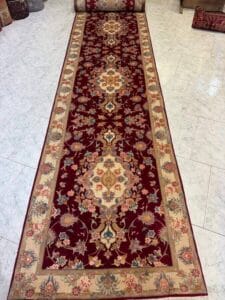 Azerbaijan Tappeti Milano Un lungo tappeto rettangolare rosso in stile persiano con intricati motivi floreali e medaglioni nei toni del crema, dell'oro e del blu, delimitati da un bordo beige. Il tappeto è esposto su un pavimento piastrellato bianco.