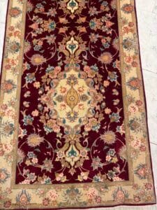 Azerbaijan Tappeti Milano Tappeto rettangolare con medaglione centrale color crema e oro, circondato da intricati motivi floreali rosa, blu e verdi su uno sfondo rosso intenso, delimitato da un disegno floreale beige; nell'angolo in alto a destra compare il logo di una foglia colorata.