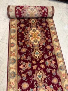 Azerbaijan Tappeti Milano Un rotolo di tappeto a motivi floreali rosso e beige è parzialmente srotolato su un pavimento piastrellato chiaro. Il tappeto presenta intricati disegni floreali e bordi ornati.