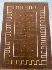 Azerbaijan Tappeti Milano Un tappeto rettangolare marrone con un bordo geometrico e piccoli motivi a stelle multicolori al centro. Un logo colorato è nell'angolo in alto a destra.