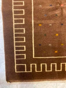 Azerbaijan Tappeti Milano Tappeto marrone con un motivo geometrico a bordo e piccoli motivi a stelle colorate. Un logo decorativo con elementi multicolori è nell'angolo in alto a destra.