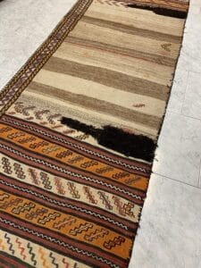 Azerbaijan Tappeti Milano Un tappeto colorato e decorato con motivi geometrici è steso su un pavimento piastrellato. Un logo multicolore a forma di foglia è visibile sull'angolo del tappeto.