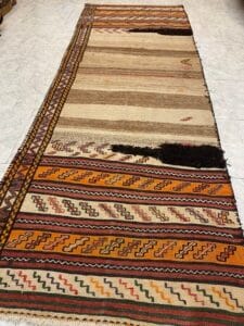 Azerbaijan Tappeti Milano Un tappeto colorato, con motivi geometrici e qualche segno di usura, adagiato su un pavimento piastrellato chiaro.