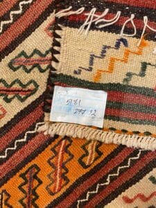 Azerbaijan Tappeti Milano Primo piano di un tessuto intrecciato con motivi geometrici in rosso, arancione e beige. Una piccola etichetta bianca con numeri scritti a mano è attaccata al tessuto.