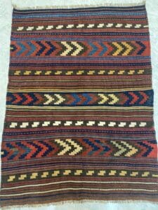 Azerbaijan Tappeti Milano Un tessuto colorato intrecciato con motivi a zigzag e geometrici nei toni del rosso, blu, beige e verde, caratterizzato da un motivo decorativo con piante nell'angolo in alto a destra.