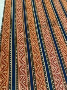 Azerbaijan Tappeti Milano Primo piano di un tessuto con motivi geometrici rossi, neri e blu navy. Un logo a farfalla colorata è nell'angolo.