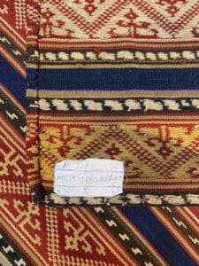 Azerbaijan Tappeti Milano Primo piano di un tessuto intrecciato con motivi geometrici in rosso, blu e beige. Un'etichetta di carta recita "ID: 107 METRE: 180 x 175". Un logo a forma di foglia colorata è presente nell'angolo in alto a destra.