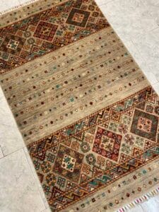 Azerbaijan Tappeti Milano Un tappeto colorato con motivi geometrici e floreali su un pavimento piastrellato. Un logo multicolore con una lettera stilizzata è nell'angolo in alto a destra.