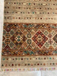 Azerbaijan Tappeti Milano Una porzione di un tappeto colorato, con motivi geometrici e un logo multicolore nell'angolo. Il tappeto è su un pavimento piastrellato.