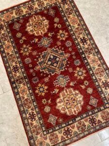 Azerbaijan Tappeti Milano Un tappeto con motivi rossi e beige, con intricati disegni geometrici e floreali disposti su un pavimento piastrellato.