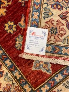 Azerbaijan Tappeti Milano Primo piano dei bordi del tappeto a motivi con etichetta "Azerbaijan" attaccata. Il tappeto presenta motivi geometrici e floreali rossi, blu e crema.