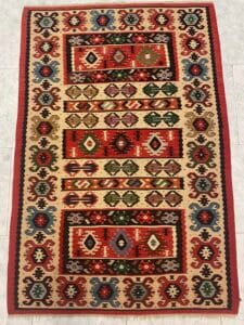 Azerbaijan Tappeti Milano Un tappeto rettangolare colorato con motivi geometrici simmetrici in rosso, verde, blu e marrone, disposto su uno sfondo chiaro e disposto su un pavimento piastrellato chiaro.