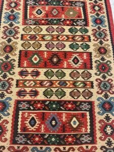 Azerbaijan Tappeti Milano Tappeto colorato con motivi geometrici a forma di rombi rossi, verdi e blu, bordato con uno sfondo marrone chiaro. Una grafica colorata a forma di farfalla è vicino all'angolo in alto a destra.