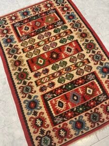 Azerbaijan Tappeti Milano Tappeto colorato con motivi geometrici nei toni del rosso, blu, verde e beige, con un emblema decorativo in un angolo, posizionato su un pavimento piastrellato.