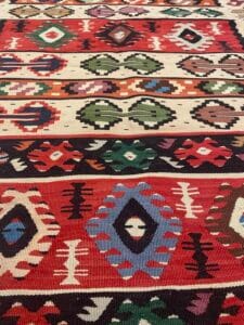 Azerbaijan Tappeti Milano Un tessuto colorato con motivi geometrici, caratterizzato da forme rosse, blu, verdi e nere su uno sfondo a strisce. Una grafica a farfalla è nell'angolo in alto a destra.