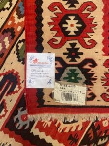 Azerbaijan Tappeti Milano Primo piano di un tessuto a fantasia con disegni geometrici in rosso, nero e beige. Sono attaccate due etichette, una con testo e l'altra con un codice a barre. È visibile un piccolo disegno ricamato.