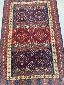 Azerbaijan Tappeti Milano Su un pavimento piastrellato di colore chiaro è esposto un tappeto con motivi geometrici e floreali nei toni del rosso, del blu e del beige.