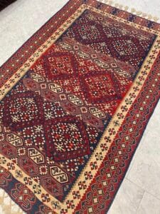 Azerbaijan Tappeti Milano Un tappeto rettangolare multicolore con motivi geometrici in rosso, blu, beige e nero su un pavimento piastrellato chiaro.