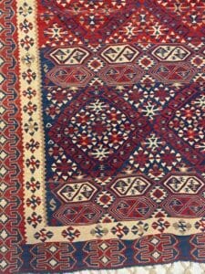 Azerbaijan Tappeti Milano Primo piano di un tappeto tradizionale con motivi geometrici in rosso, blu e beige, con una piccola farfalla nell'angolo in alto a destra.