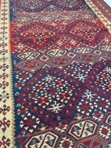 Azerbaijan Tappeti Milano È esposto un tappeto con un motivo geometrico rosso, blu e crema. Un logo a forma di stella colorata con il testo "Aladdin Rugs" è nell'angolo.
