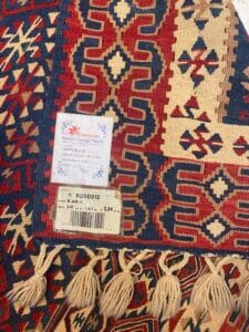 Azerbaijan Tappeti Milano Tessuto con motivi intricati con disegni geometrici e frange. Sono visibili due etichette, una con testo e l'altra con un logo raffigurante una foglia colorata.